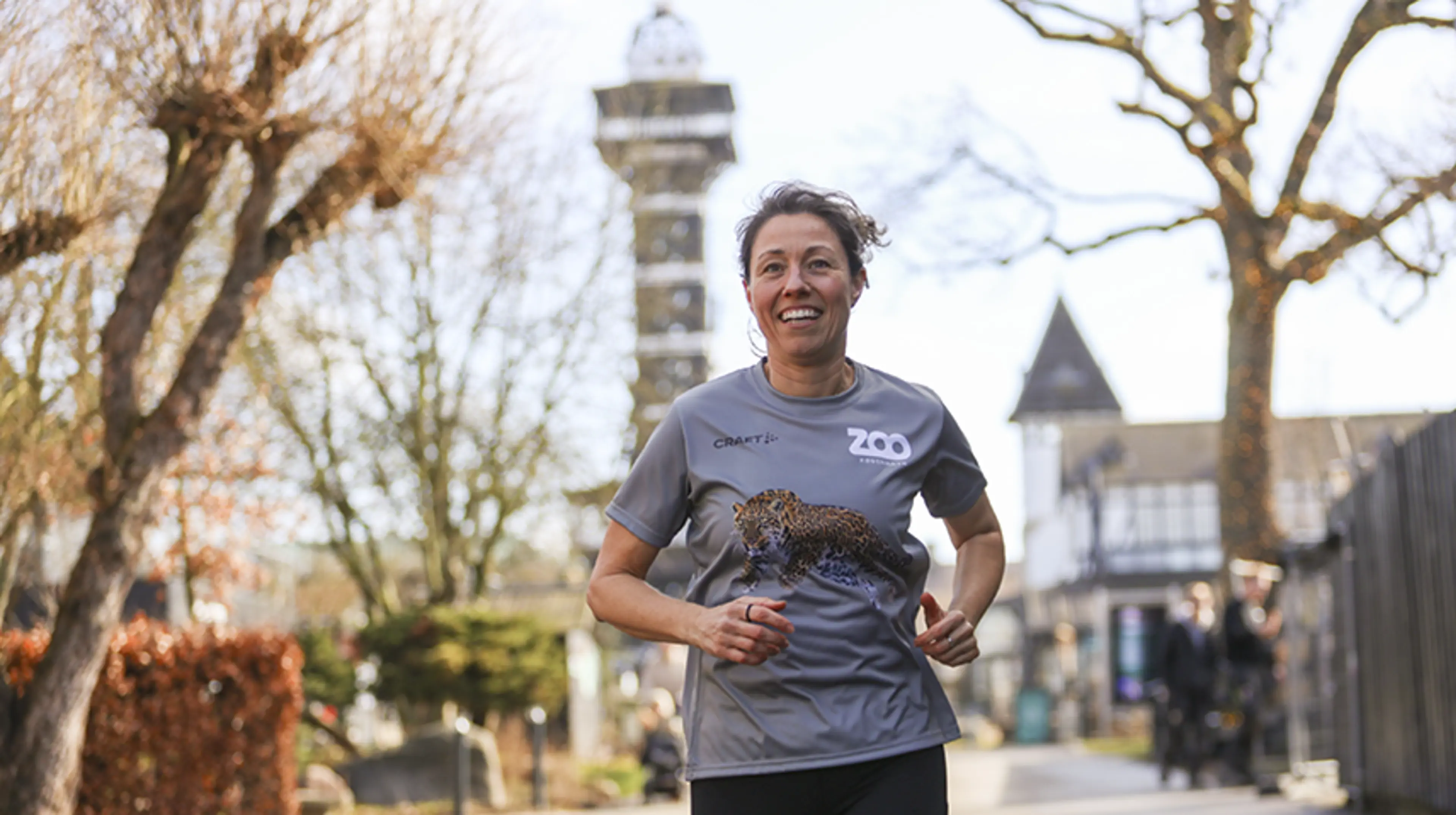Kvindelig løber i grå løbetrøje med ZOO København-logo smiler under løb i ZOO med Tårnet bag sig